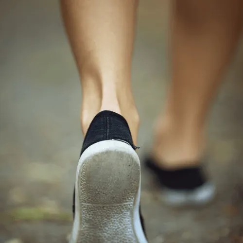 Close-up of person walking on pavement wearing black slip-on shoes with white soles.