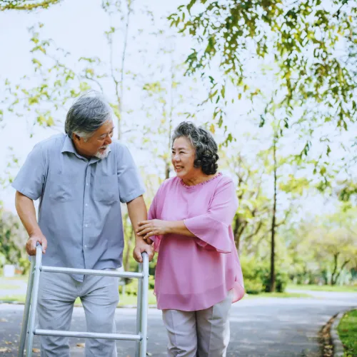 Elderly man using a walker supported by a smiling woman in a pink blouse walking outdoors in a park.