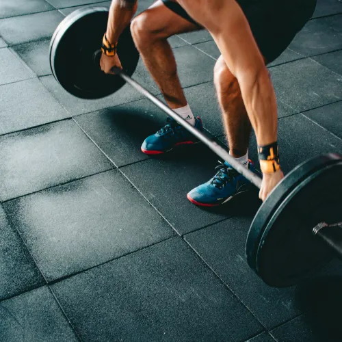Athlete lifting a barbell in a gym with rubber flooring, wearing blue shoes and wrist wraps.