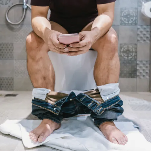 Man sitting on toilet with pants down, using smartphone in modern tiled bathroom.