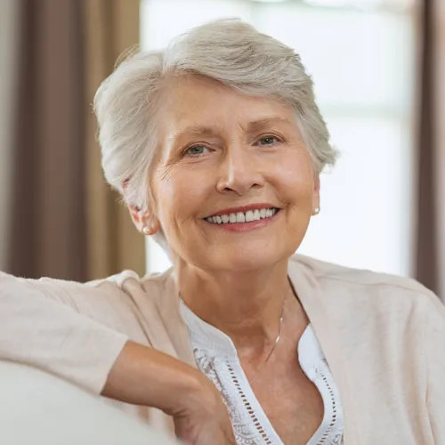 Smiling elderly woman with short gray hair sitting comfortably indoors, wearing a light cardigan and white blouse.