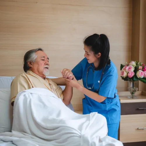Nurse in blue scrubs comforting elderly male patient in bed with white blanket in cozy room.