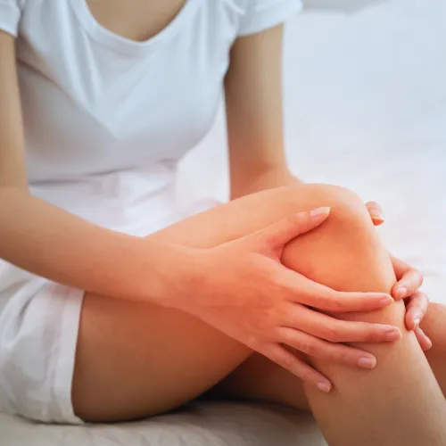 Woman sitting on bed holding her knee, indicating knee pain or discomfort, wearing white clothing.