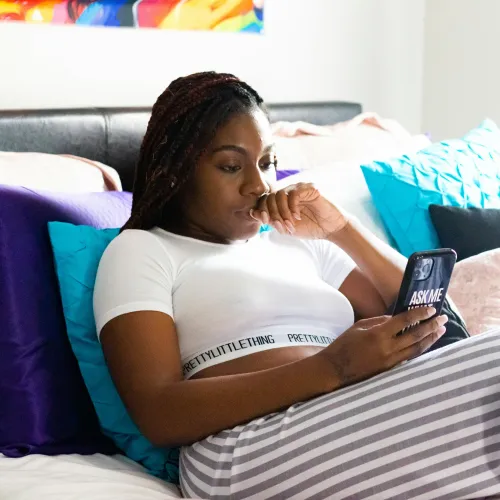 Young woman in striped pants using smartphone while relaxing on a bed with colorful pillows in bright room.