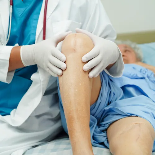 Doctor examining an elderly patient's knee with visible surgical scars in a hospital room setting.
