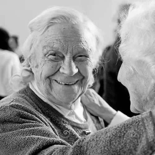 Two elderly women smiling warmly and embracing each other in a heartfelt moment in black and white.
