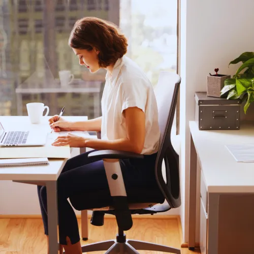 Young woman working at a desk with laptop and notebook in a bright office space near a window.