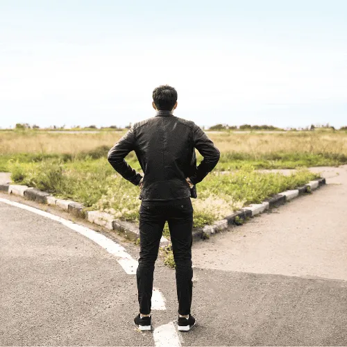 Man in black jacket standing at a forked road in a rural area facing two divergent paths.