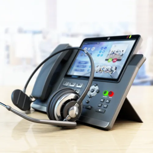 Modern office desk phone with touchscreen display and headset on a wooden surface in bright workspace.