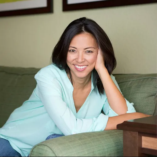 Smiling woman with dark hair wearing a light blue blouse sitting comfortably on a green couch indoors.