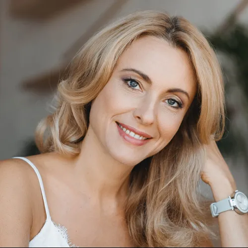 Smiling woman with long blonde hair, wearing a white top and a wristwatch, sitting indoors.
