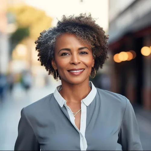 Smiling middle-aged woman with curly gray hair wearing hoop earrings and a gray blouse outdoors with blurred background