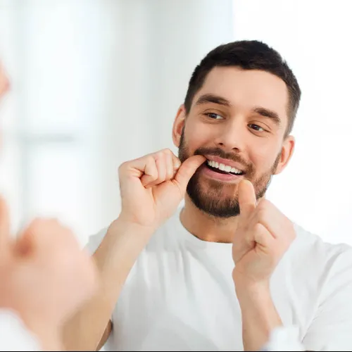 Young man smiling while flossing his teeth in front of a mirror in a bright bathroom.