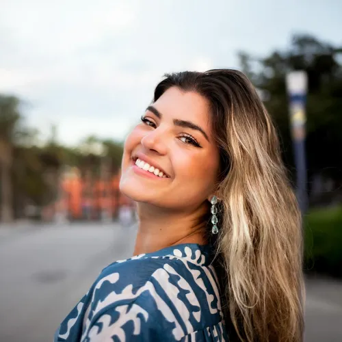 Smiling woman with long hair and earrings, wearing a blue patterned top, outdoors with soft background.