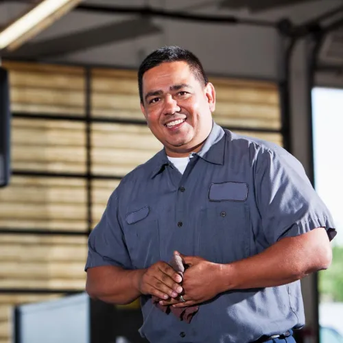 Smiling mechanic in work uniform stands confidently in a garage environment, showcasing teamwork and professionalism.
