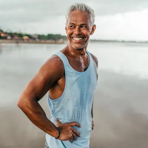 Smiling older man in a tank top standing on the beach at sunset with reflection in the water.