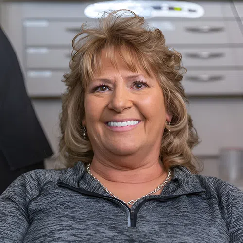 Smiling middle-aged woman with curly blonde hair wearing gray zip-up sweater in an office setting.