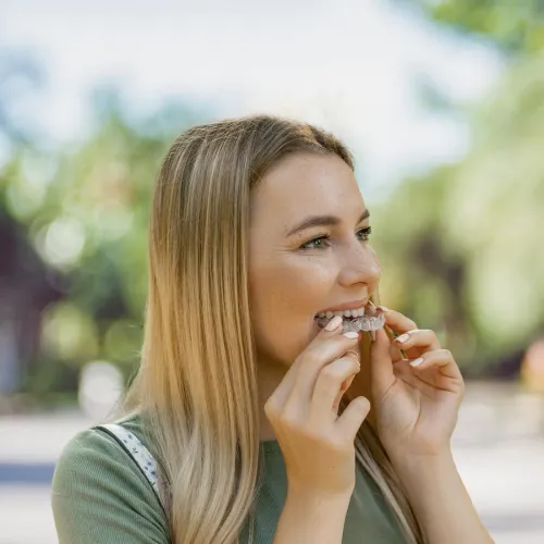Woman with long hair holding teeth aligners outdoors, smiling in natural light.