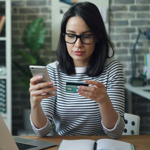 Woman wearing glasses is shopping online using phone and credit card at desk with laptop and notebook.