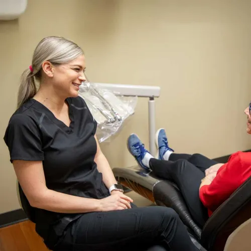 Dentist in black scrubs smiling and talking with senior patient seated in dental chair