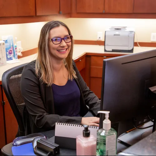 Smiling woman with glasses working at a computer in a modern office with wood cabinets and hand sanitizer bottles.