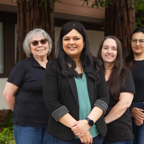 Group of four diverse women standing outdoors smiling in casual and professional attire near trees and greenery