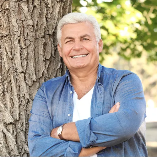 Smiling elderly man with white hair leaning against a tree outdoors wearing a denim shirt and white tee.