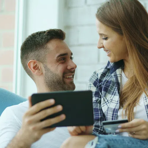 Couple shopping online with tablet and credit card, smiling and sitting together in a bright room.