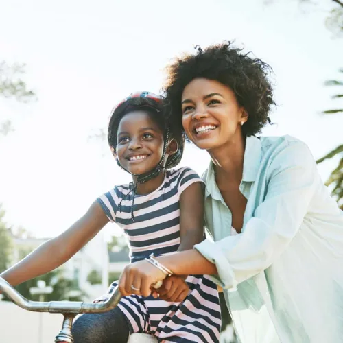 A joyful mother and daughter smiling together while riding a bicycle outdoors in sunny weather.