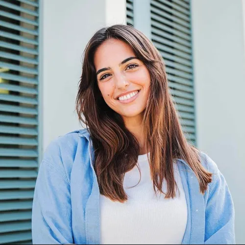 Smiling young woman with long brown hair wearing a white top and light blue shirt standing outdoors.