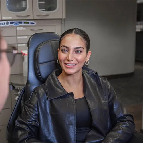 Smiling woman in black leather jacket sitting and talking to another person in an indoor office setting