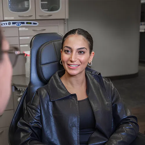 Smiling woman in black leather jacket sitting in a dental chair talking to a dentist in clinic.