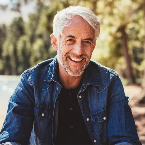 Smiling mature man with gray hair and beard wearing denim jacket outdoors with blurred forest background.
