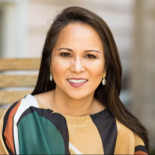 Smiling middle-aged woman with long dark hair wearing a colorful patterned blouse and white earrings.