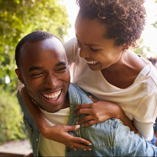 Smiling couple enjoying a piggyback ride outdoors with greenery in the background on a sunny day