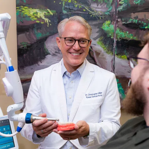 Smiling male dentist in white coat demonstrating dental tool on teeth model to patient in clinic.