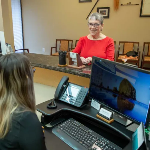 Older woman in red shirt smiling at receptionist across the counter in a professional office setting.