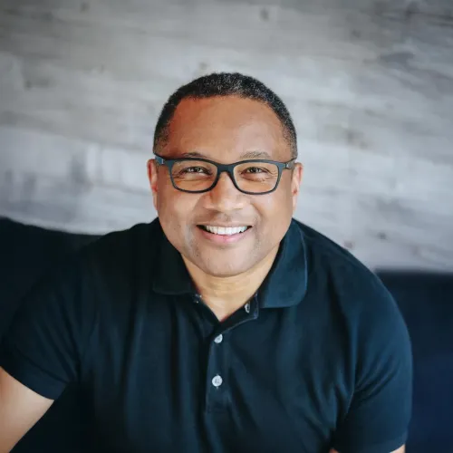 Smiling man with glasses wearing a black polo shirt against a light gray wooden background