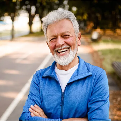 Smiling elderly man with white beard in blue jacket standing outdoors on a sunny day with arms crossed