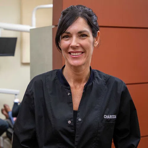 Smiling female dental professional in black scrubs in a dental office with a colleague in the background
