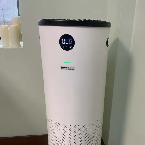 Modern cylindrical air purifier with digital display placed on wooden floor near corner wall.