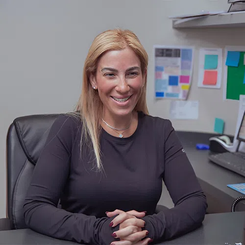 Smiling woman in black shirt sitting at desk facing another person in office with computer and notes.