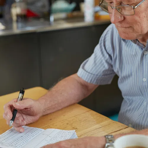 Elderly man wearing glasses writing in a notebook with numbers at a wooden table with coffee cup.