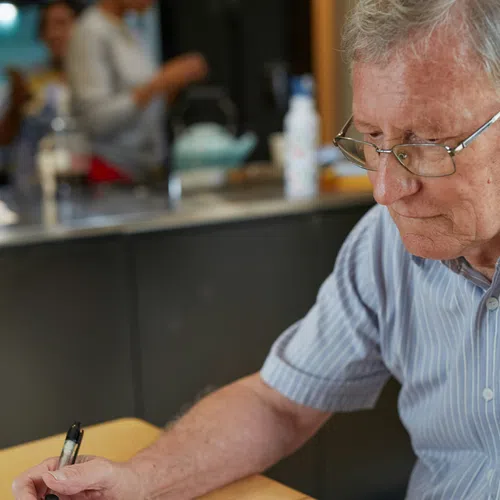 Elderly man wearing glasses writing in a notebook with numbers at a wooden table with coffee cup.