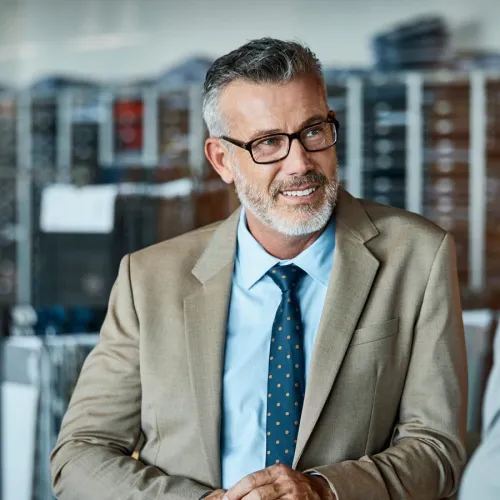 Businessman in suit and glasses smiling in modern office setting with shelves in background.