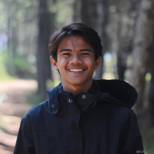 Smiling young man in a park with trees in the background, wearing a black jacket.