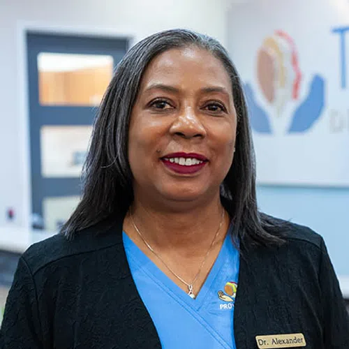 Professional woman in blue scrubs and black jacket smiling with a name tag reading Dr. Alexander indoors.