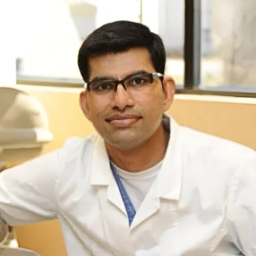 Male dentist wearing glasses and white coat leaning on dental chair with a window in the background