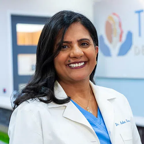 Smiling female doctor with long black hair in white coat and blue scrubs standing in medical office.