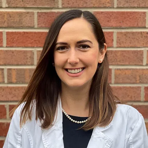 Smiling woman wearing a white lab coat, pearl necklace, black top, standing in front of a brick wall